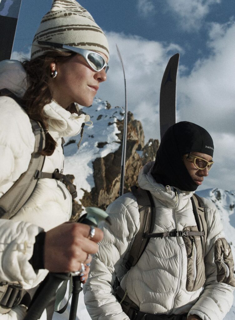 Two people in insulated ski apparel and backpacks standing in a snowy mountain landscape with skis and ski poles.