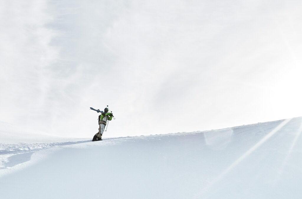 Skier hiking uphill through deep snow, carrying skis on shoulder.