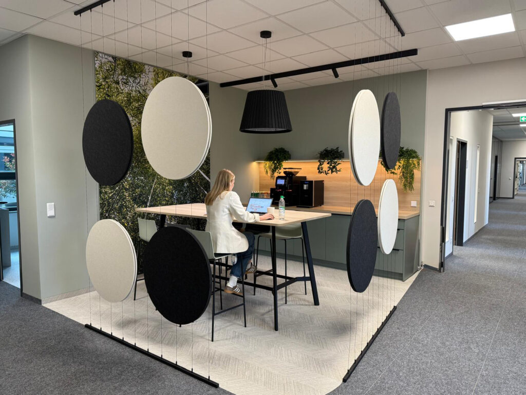 Modern office coffee area with a high table and stools, acoustic circle panels as a divider, and a person working on a laptop; background features a coffee machine, wooden backsplash, and green plants.