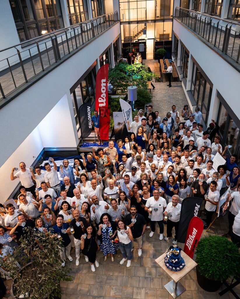 Large group of people gathered in an indoor atrium for a company event, standing around a central area with branded flags and a decorated cake on a table; the space features two levels with railings, glass doors, and greenery in the background.