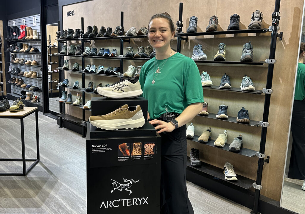 Person in a green shirt standing in a shoe store beside a display of Arc’teryx hiking shoes.