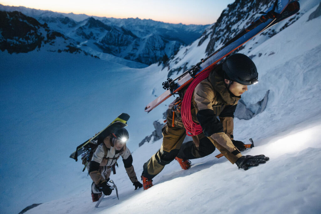 Two climbers with skis and headlamps ascending a steep snowy mountain slope at dawn.
