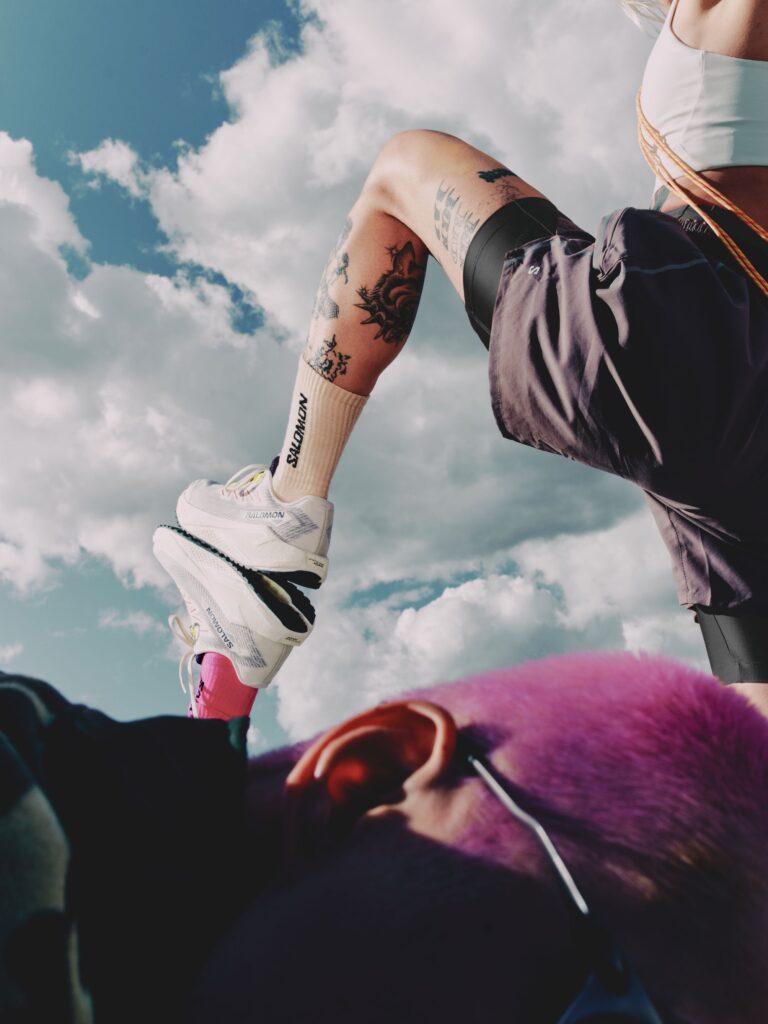 Close-up of a person’s leg wearing white Salomon sneakers and branded socks, leaning on another person’s foot with pink hair, against a bright blue sky with scattered clouds.