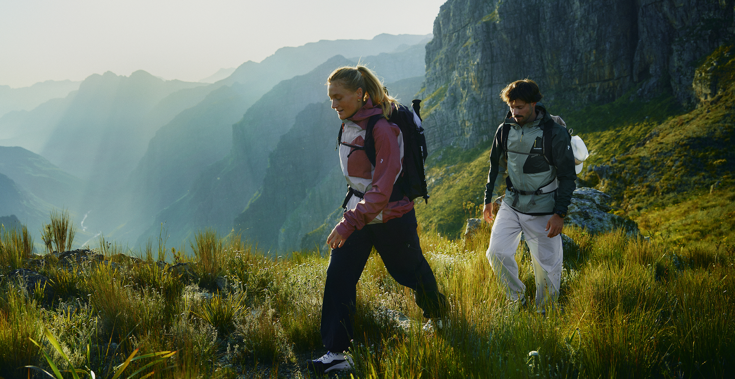 Two hikers walking along a grassy mountain trail with dramatic cliffs and sunlight in the background.