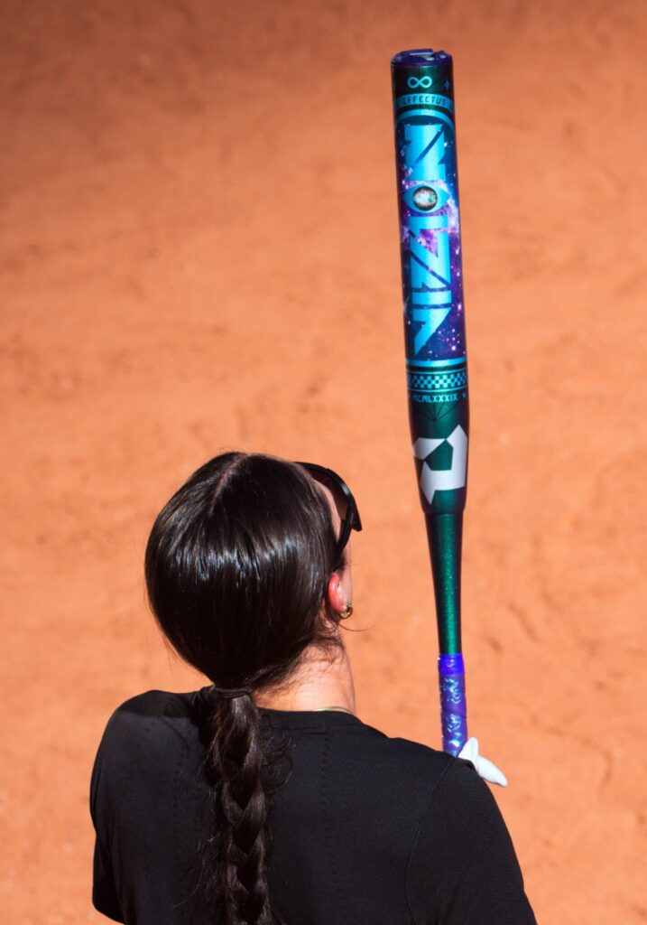 Athlete balancing a DeMarini Vizion bat vertically on a dirt field.