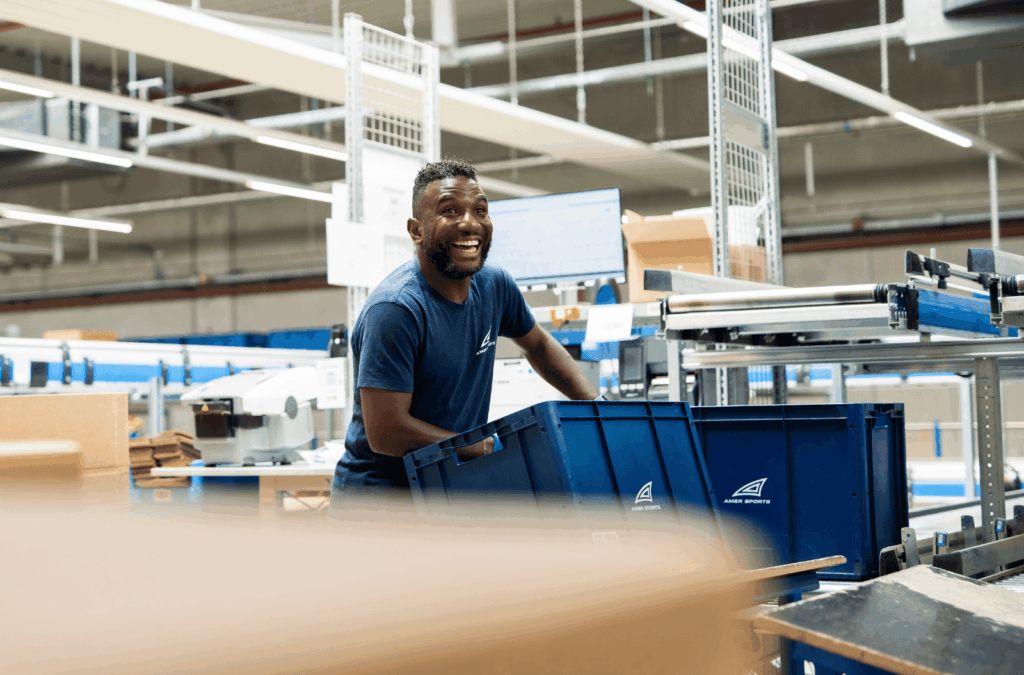 Smiling Amer Sports employee moving a blue crate in the Überherrn distribution center.